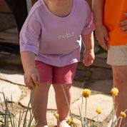 A child wearing a lavender t-shirt with the word 'cups' printed on the chest and pink shorts, standing outdoors in a garden next to another child in an orange shirt.