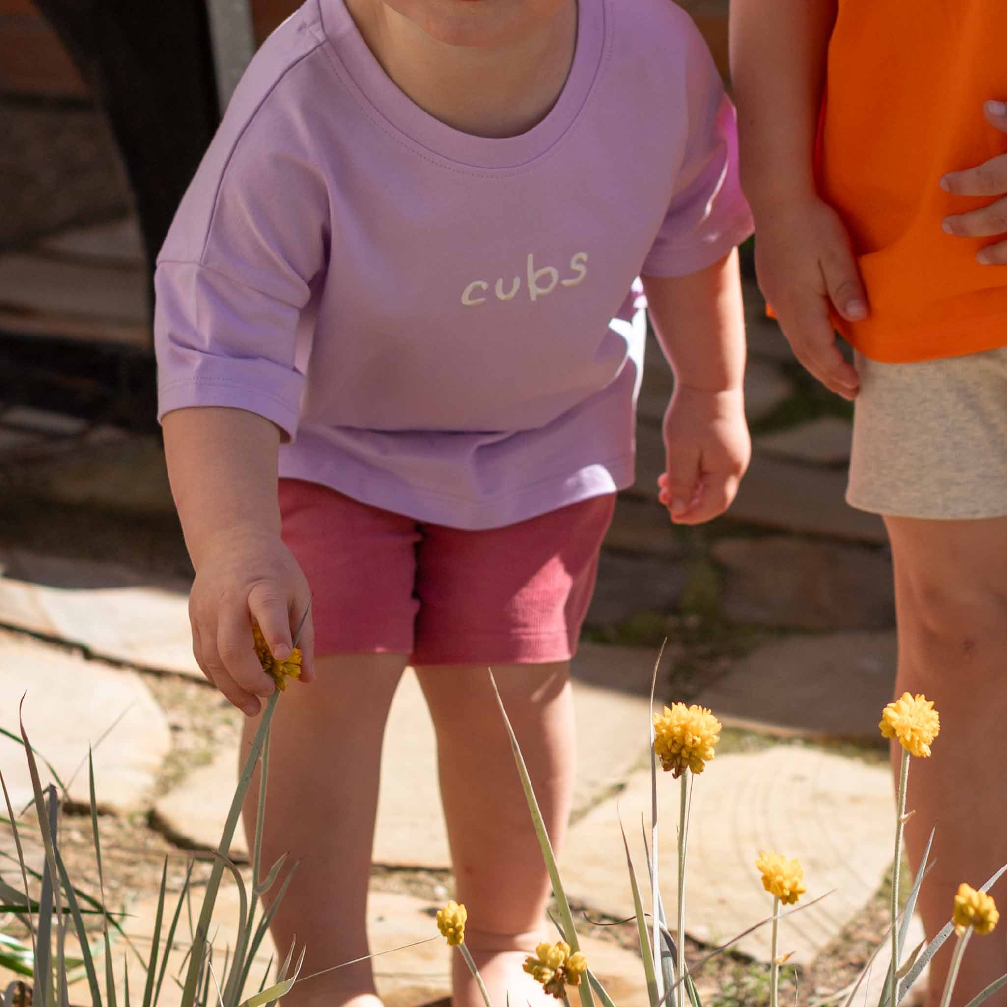 A child wearing a lavender t-shirt with the word 'cups' printed on the chest and pink shorts, standing outdoors in a garden next to another child in an orange shirt.