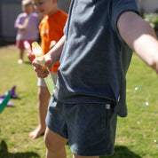 A child wearing a dark slate blue heathered t-shirt and matching shorts stands outdoors on a grassy lawn, holding a bubble wand. The background shows other children and bubbles in a sunny backyard setting.