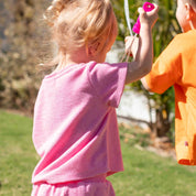 A young child seen from behind wearing a pink short-sleeve t-shirt and matching pink bottoms, standing outdoors on grass and holding a bright pink object. Another child in an orange shirt is partially visible at the right edge.