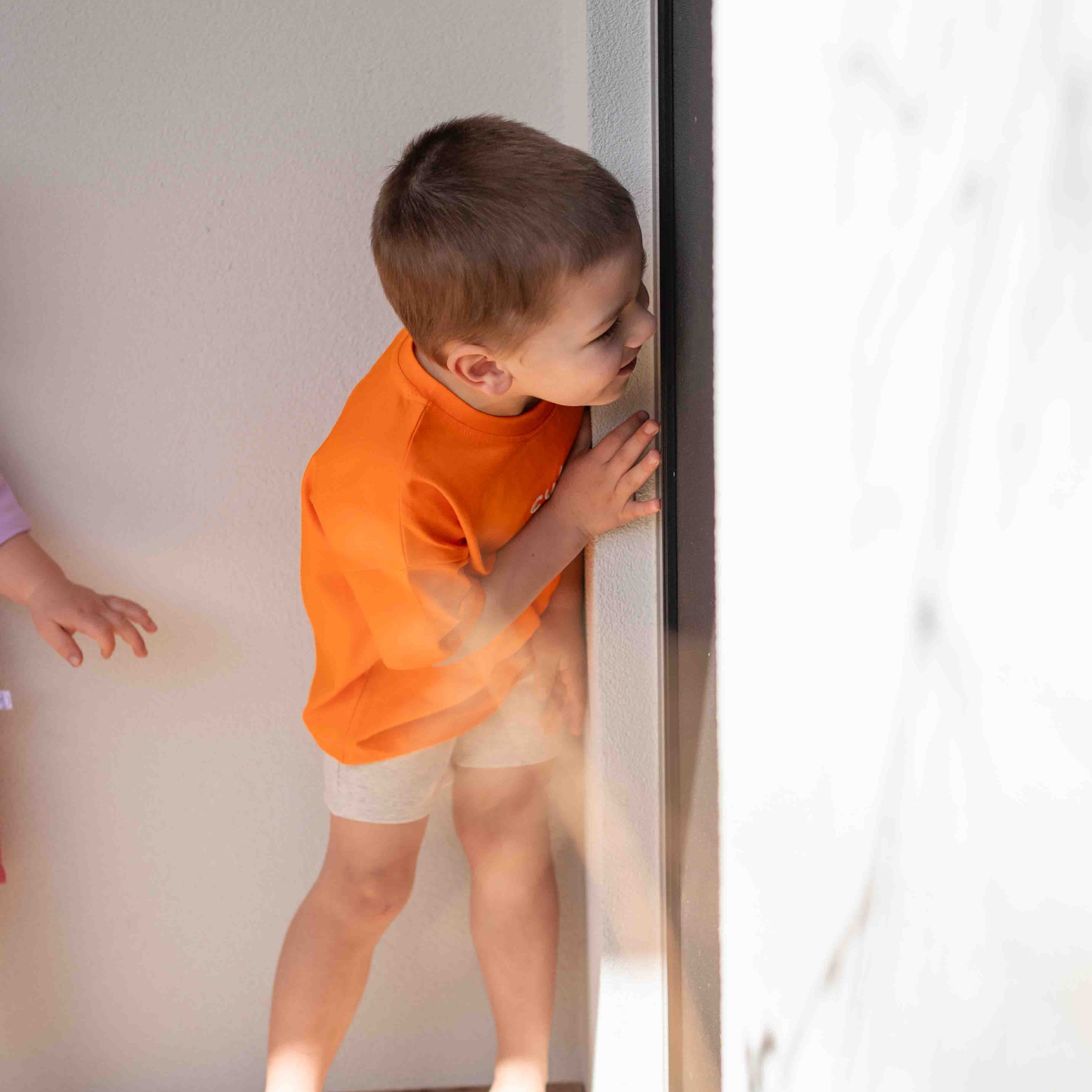 child peering around a wall corner in an orange shirt and grey bike shorts
