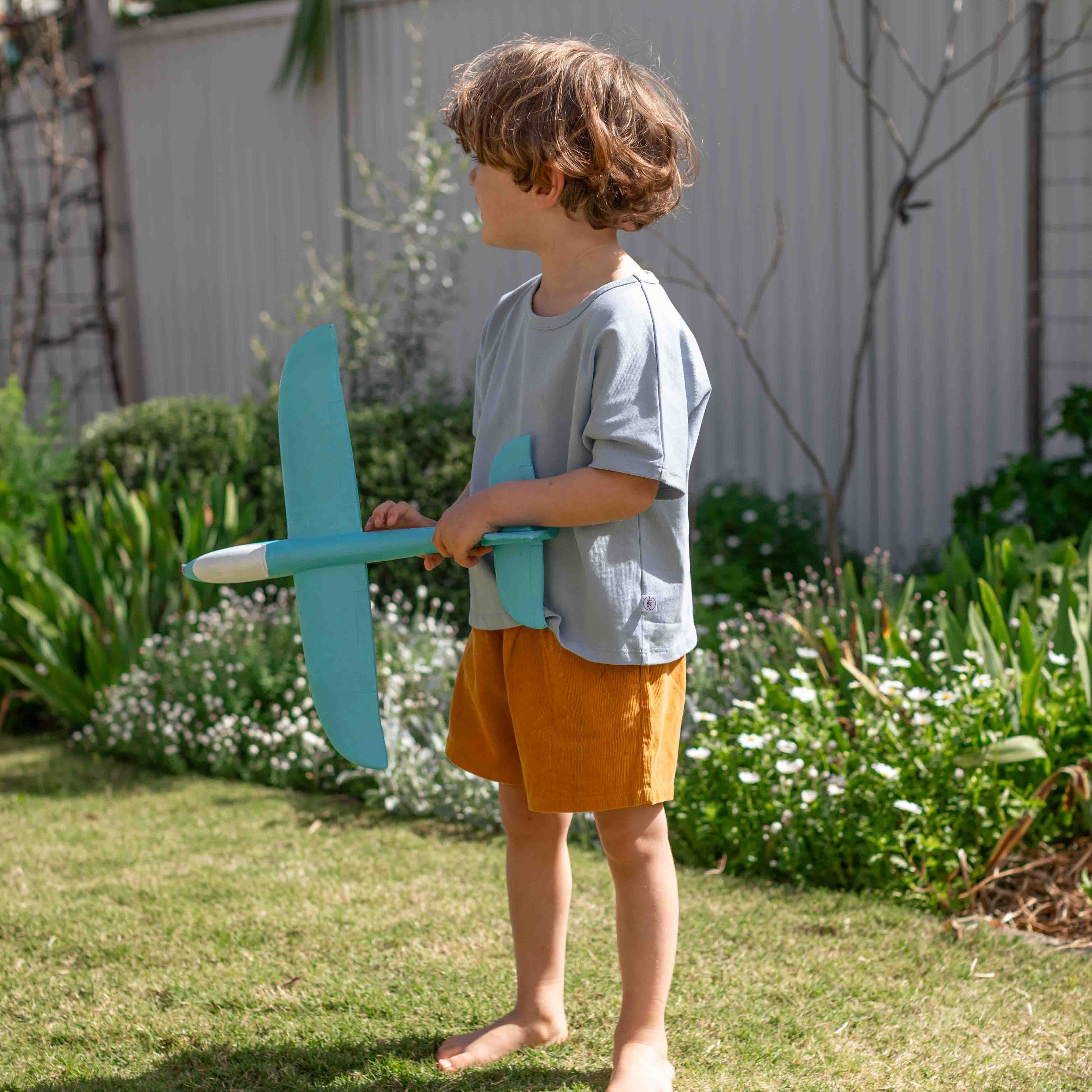 A young child with curly brown hair stands barefoot on a grassy backyard lawn, holding a light blue foam airplane. The child is wearing a light blue t-shirt and mustard yellow shorts, with a white fence and green garden plants in the background.