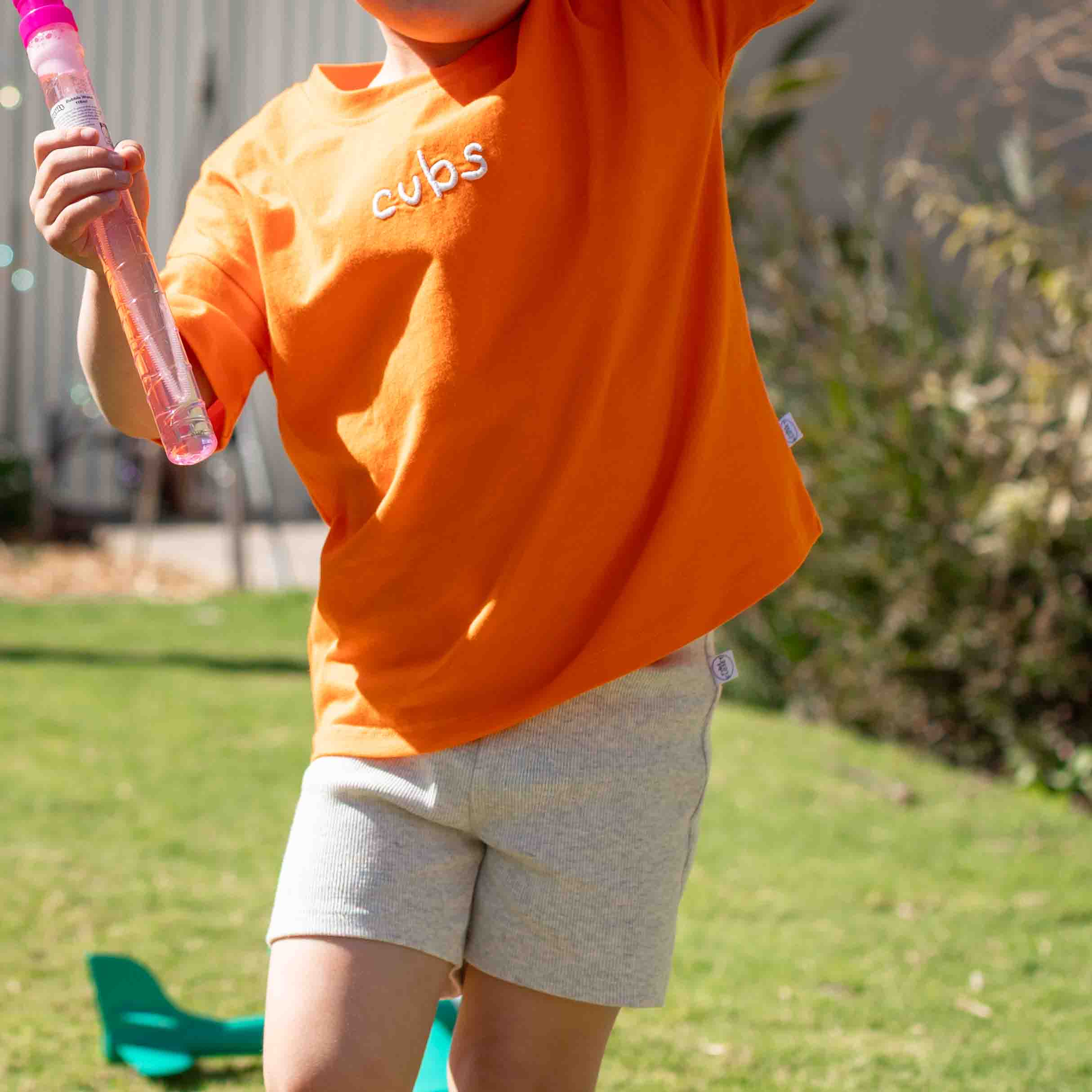 A child wearing a bright orange short-sleeve t-shirt with 'cubs' embroidered in white on the chest, paired with light grey shorts, standing outdoors on a grassy lawn.