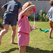 A toddler in a pink short-sleeve top and matching shorts stands on a grassy backyard playing with a bubble wand, with other children blurred in the background.