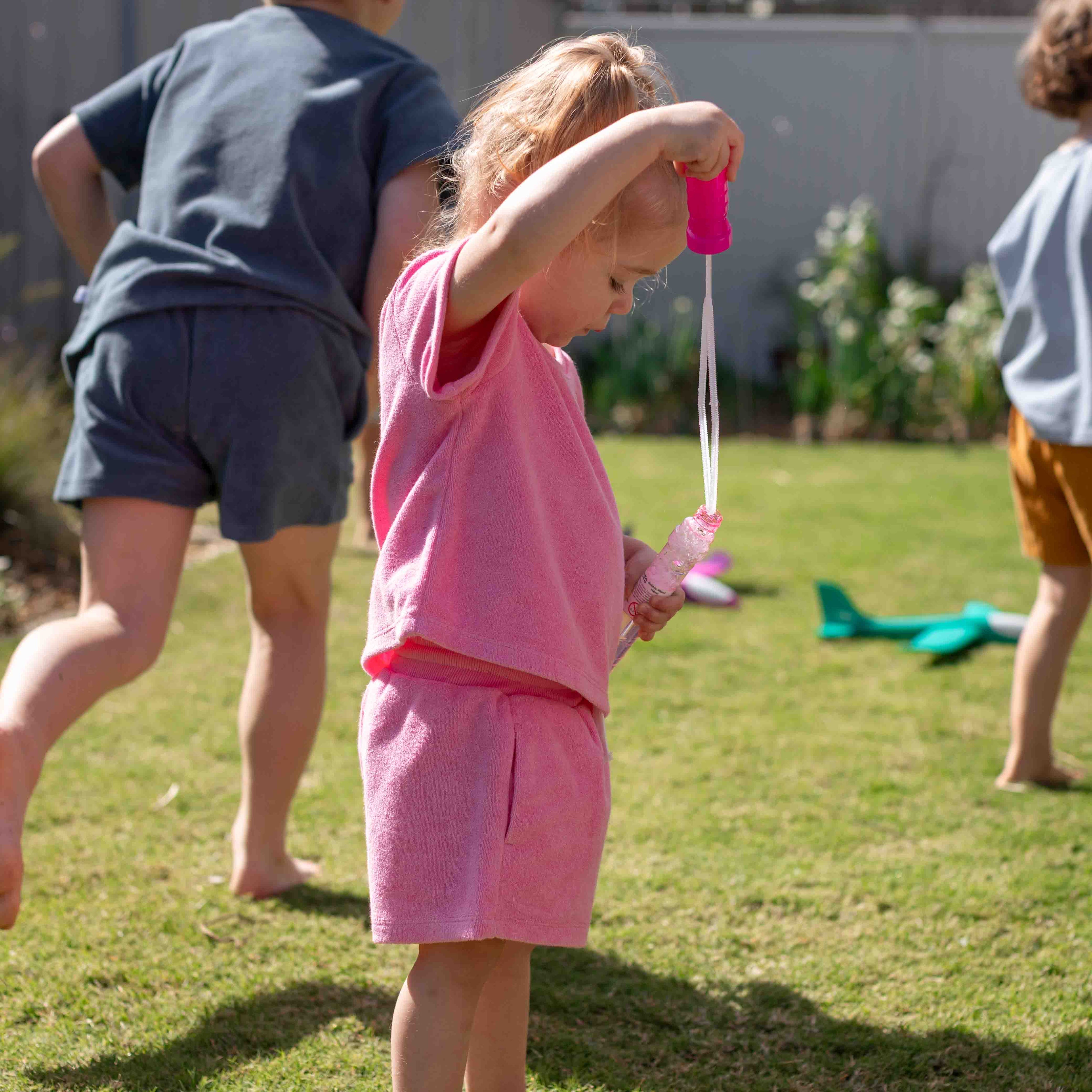 A toddler in a pink short-sleeve top and matching shorts stands on a grassy backyard playing with a bubble wand, with other children blurred in the background.