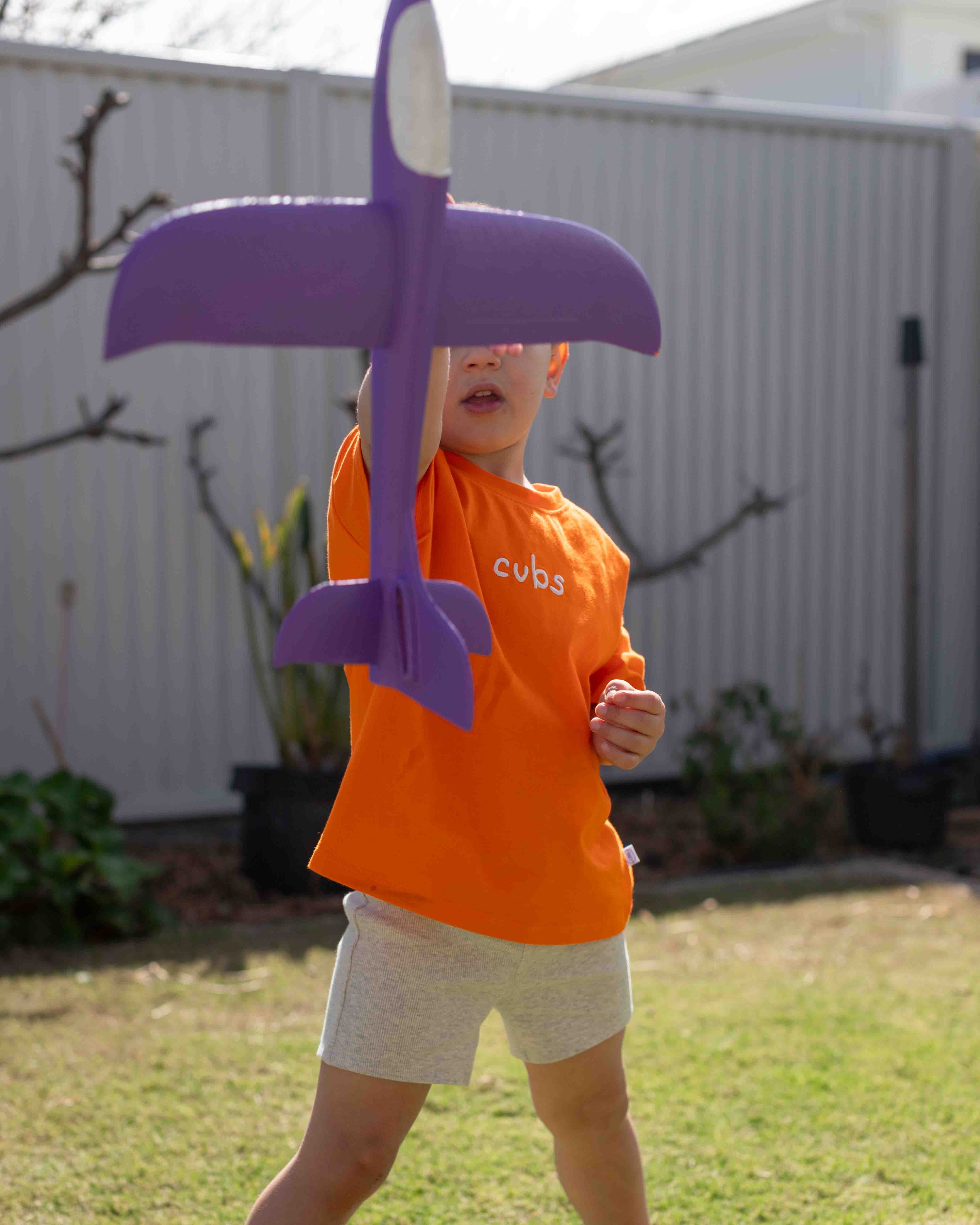 A child wearing an orange t-shirt with 'cubs' printed on it and light gray shorts, holding a large purple foam airplane in a backyard setting.