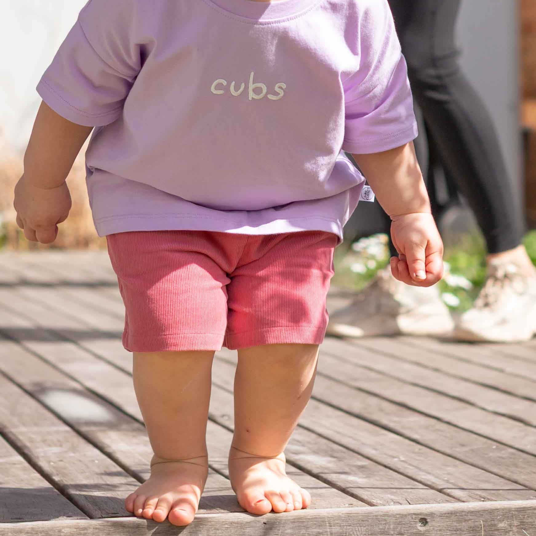 A toddler walking barefoot on a wooden deck, wearing a light purple short-sleeve t-shirt with the word 'cubs' printed in white on the chest, and pink shorts.