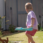 A toddler in a light purple t-shirt and pink shorts walks barefoot in a backyard while holding a turquoise and white toy airplane.