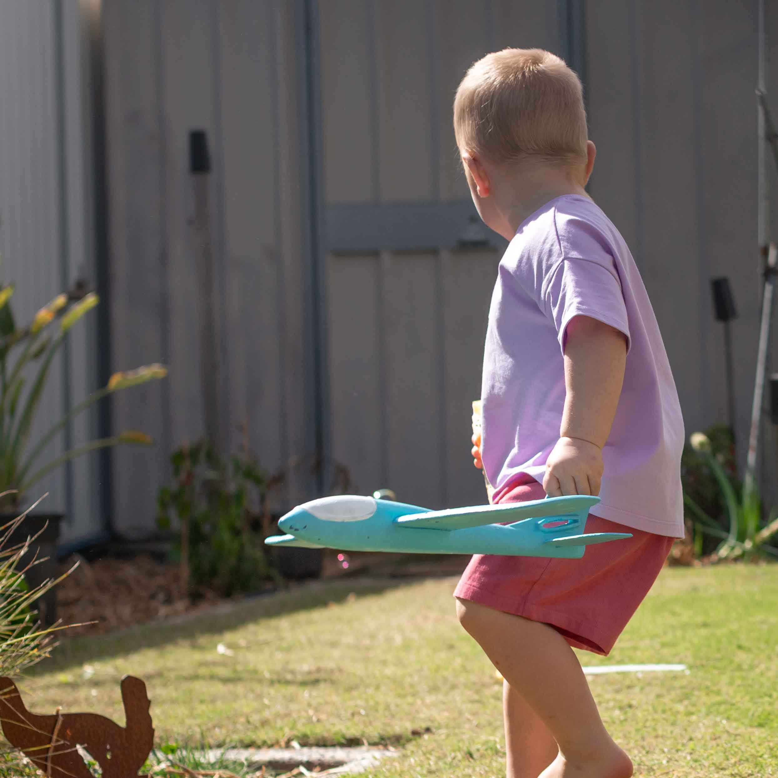 A toddler in a light purple t-shirt and pink shorts walks barefoot in a backyard while holding a turquoise and white toy airplane.