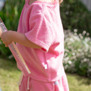 A child wearing a pink short-sleeve knit top and matching pink bottoms, standing outdoors in a garden while holding a pink toy bat.