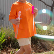 A child wearing an oversized bright orange t-shirt with the word 'cups' printed in white on the chest, holding bubble wands outdoors in a garden setting.