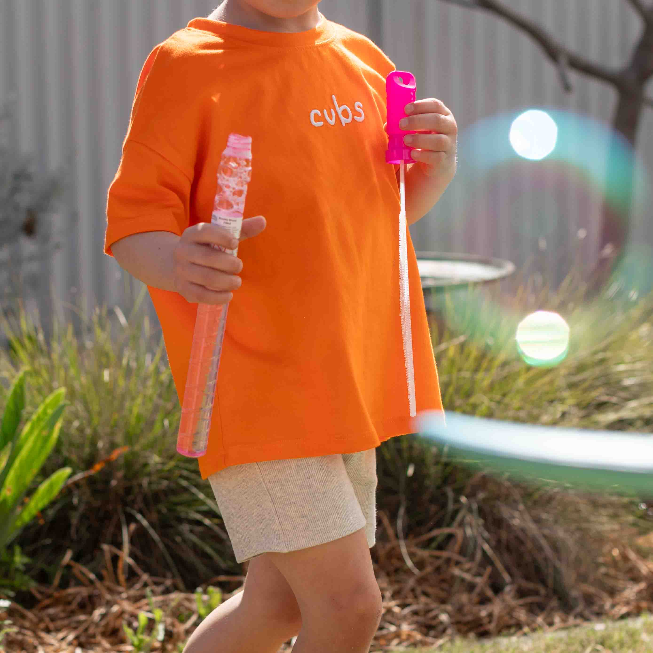 A child wearing an oversized bright orange t-shirt with the word 'cups' printed in white on the chest, holding bubble wands outdoors in a garden setting.