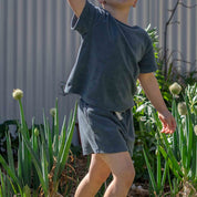 A child wearing a dark slate blue short-sleeve t-shirt and matching shorts, standing in a garden among green plants in front of a corrugated metal wall.