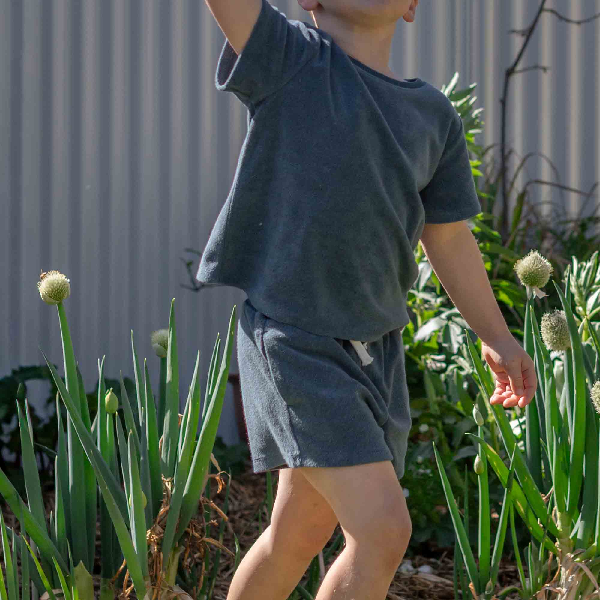 A child wearing a dark slate blue short-sleeve t-shirt and matching shorts, standing in a garden among green plants in front of a corrugated metal wall.