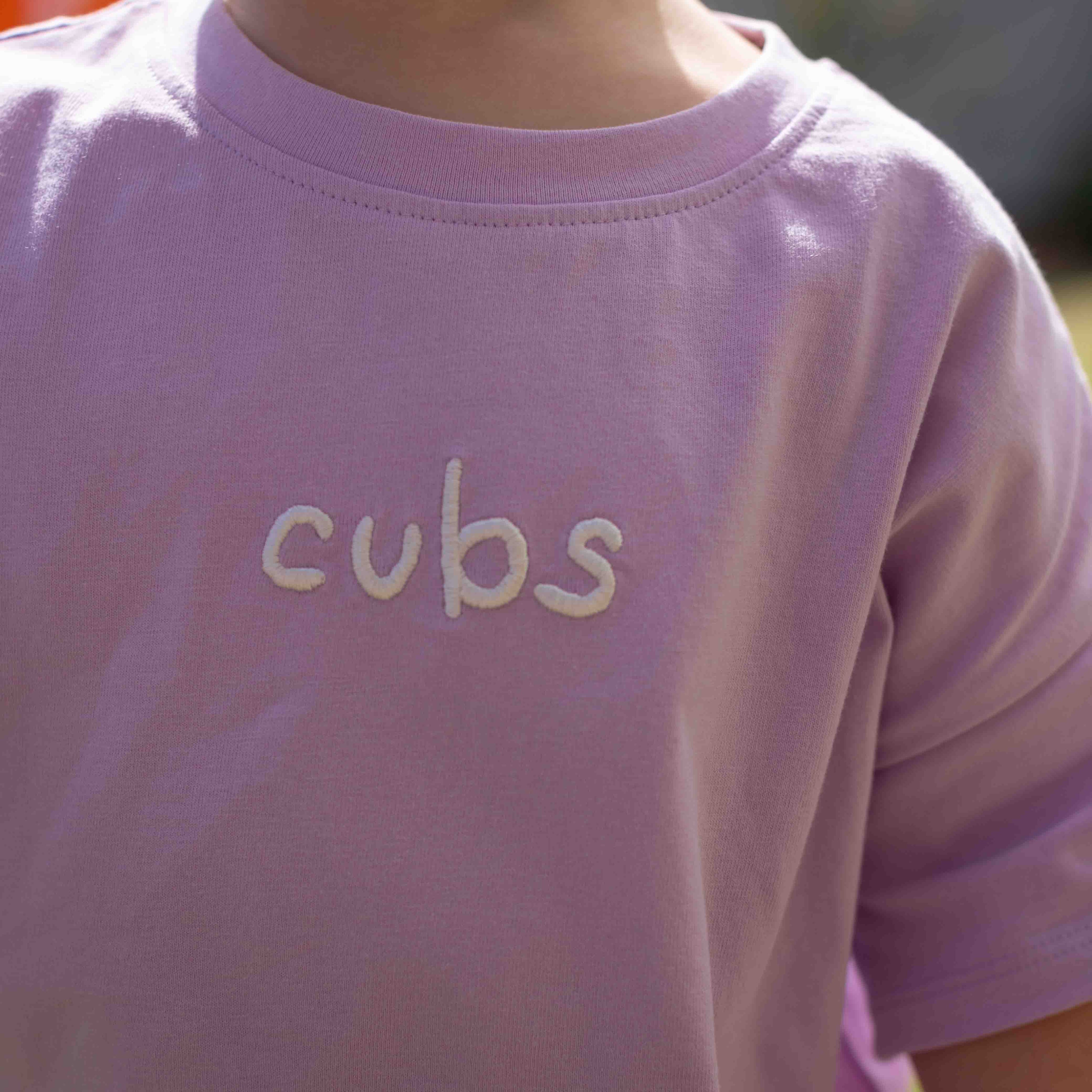 A close-up of a person wearing a light lavender crewneck t-shirt with the word 'cubs' embroidered in white across the chest.