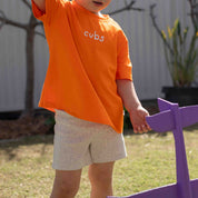 A toddler wearing a bright orange short-sleeve t-shirt with the word 'cubs' printed in white on the chest, paired with light heather gray shorts, standing outdoors in a backyard next to a purple toy.