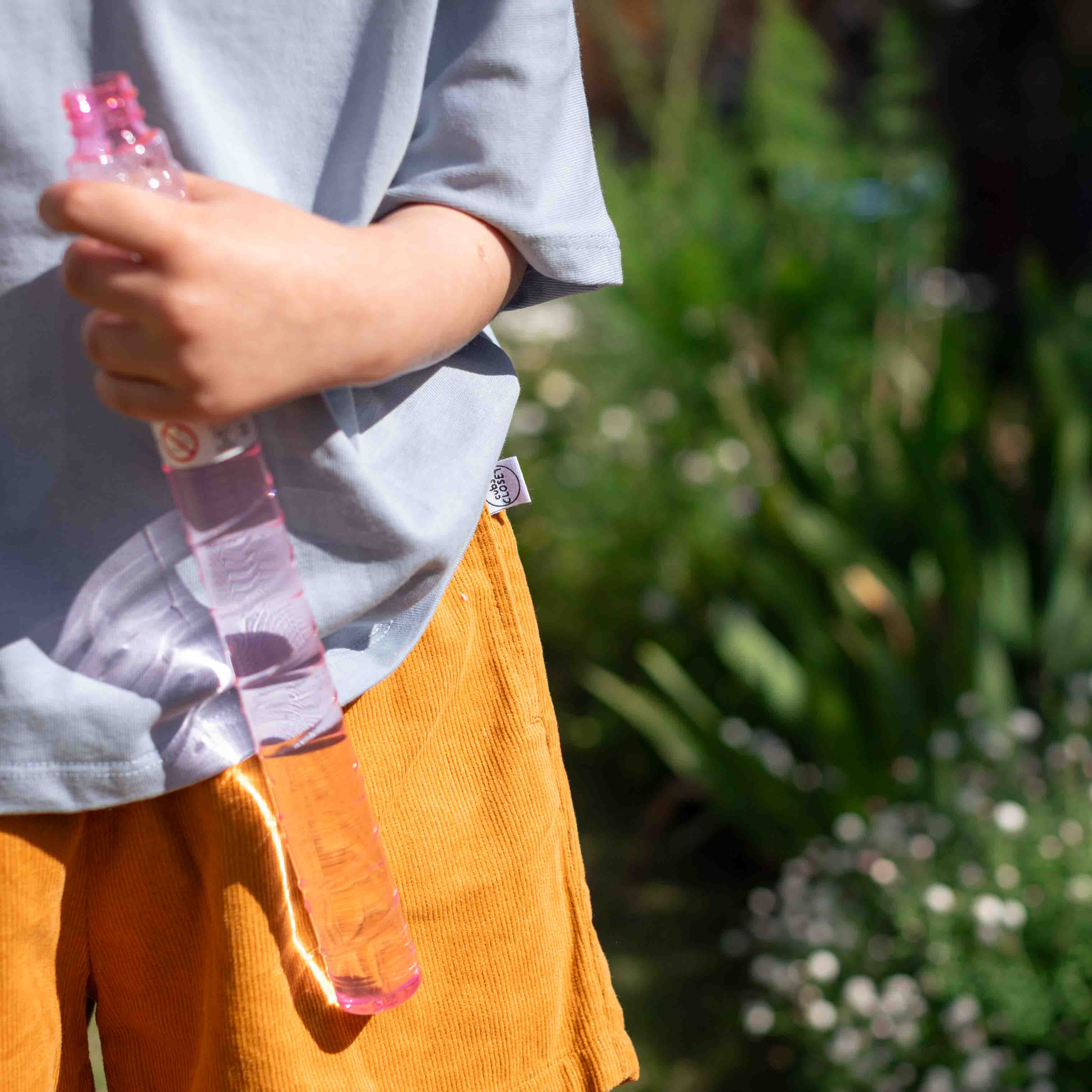 A child outdoors wearing a light blue t-shirt and orange corduroy shorts, holding a clear plastic bottle with pink liquid and a pink cap. The background is a soft-focus green garden.