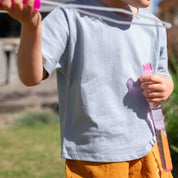A child wearing a light blue short-sleeve t-shirt and orange pants, holding a pink bubble wand outdoors in a sunny yard.