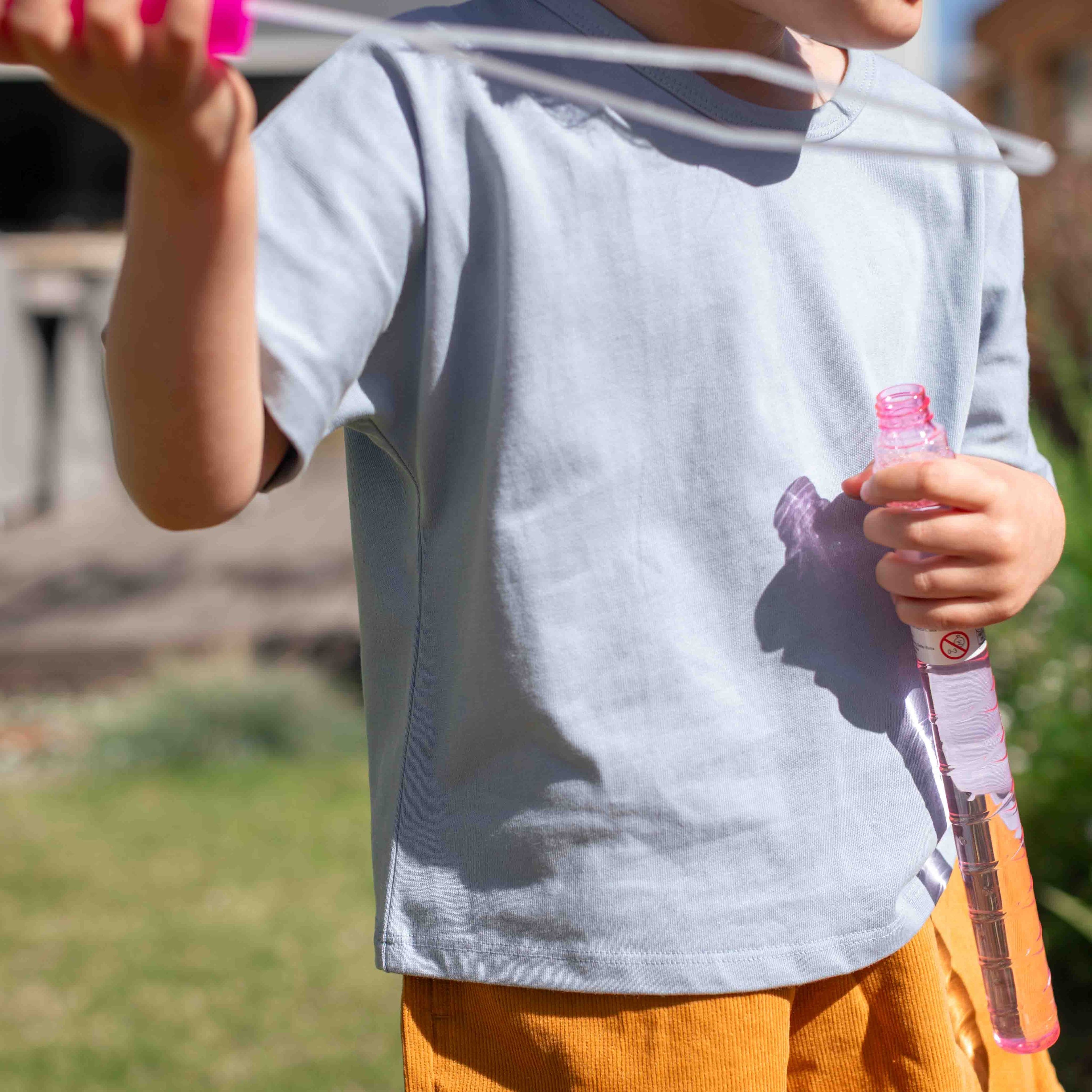 A child wearing a light blue short-sleeve t-shirt and orange pants, holding a pink bubble wand outdoors in a sunny yard.