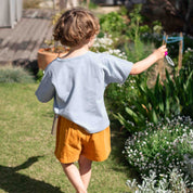 A child seen from behind wearing a light blue short-sleeve t-shirt and mustard yellow shorts, walking in a sunny garden.