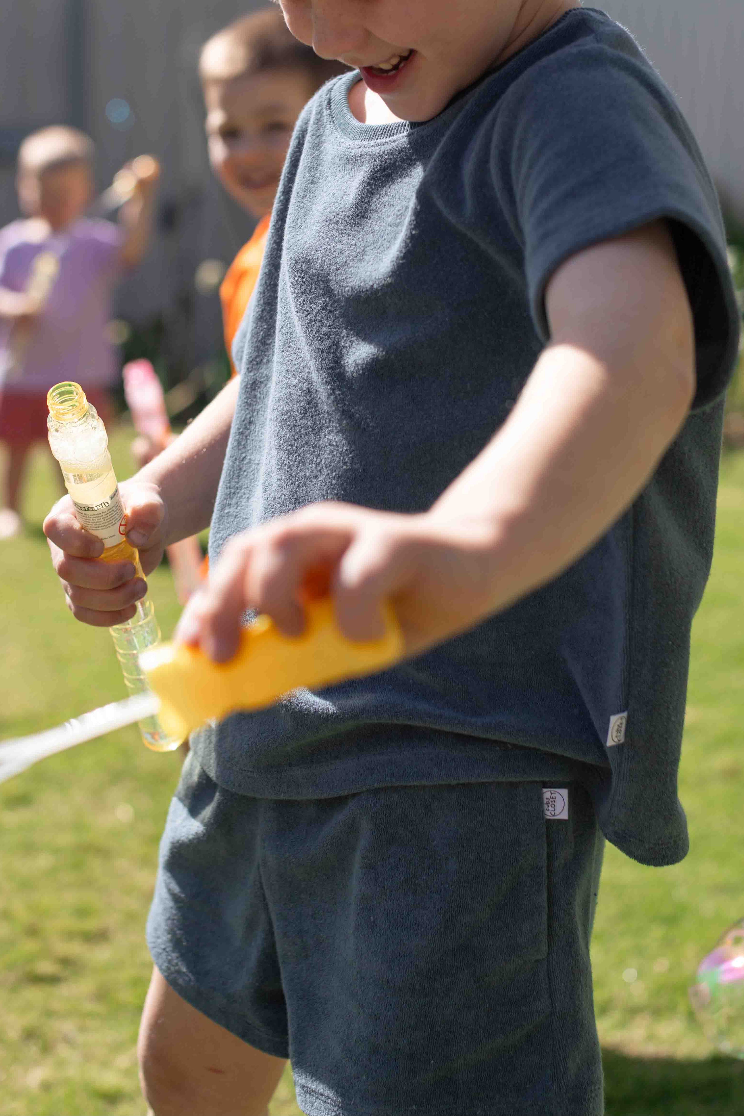 A young boy in a navy blue t-shirt and matching shorts smiles while pouring bubble solution from a yellow bottle into a bubble wand in an outdoor backyard setting.