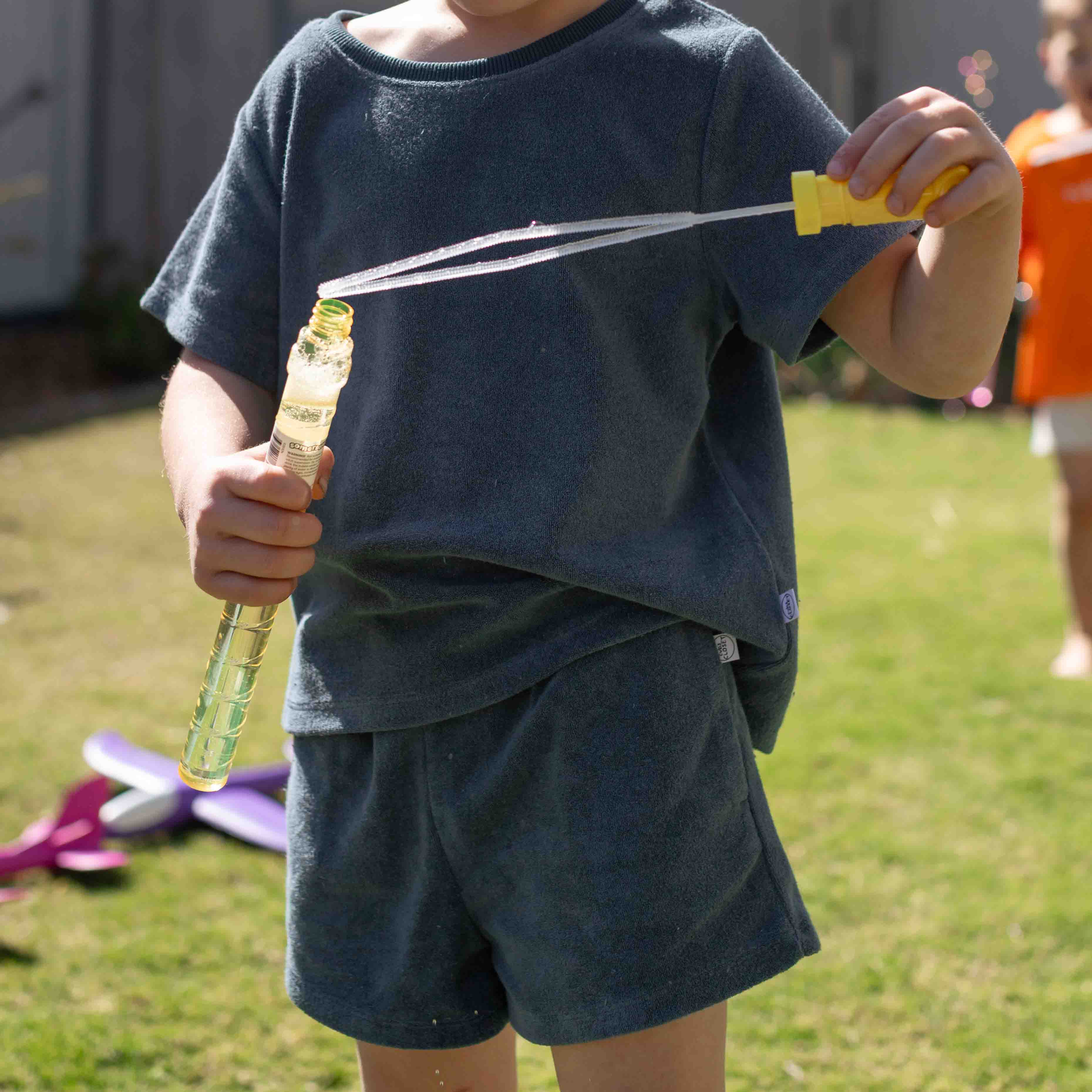 A child wearing a dark slate blue t-shirt and matching shorts is outdoors on a grassy lawn, holding a bubble wand with a yellow handle.