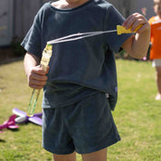 A child wearing a dark slate blue t-shirt and matching shorts is outdoors on a grassy lawn, holding a bubble wand with a yellow handle.