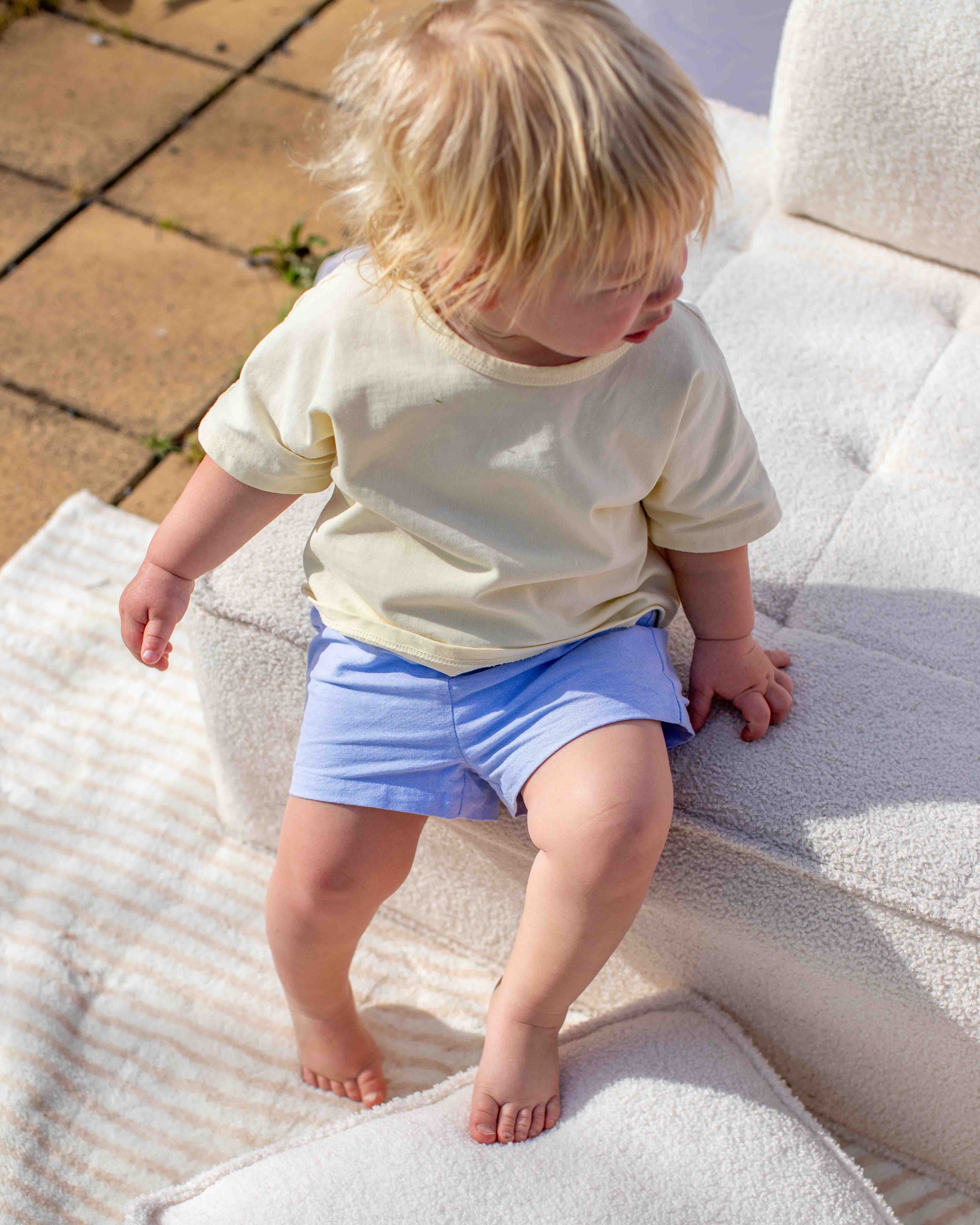 Child wearing a yellow shirt and blue shorts sitting on a textured surface.