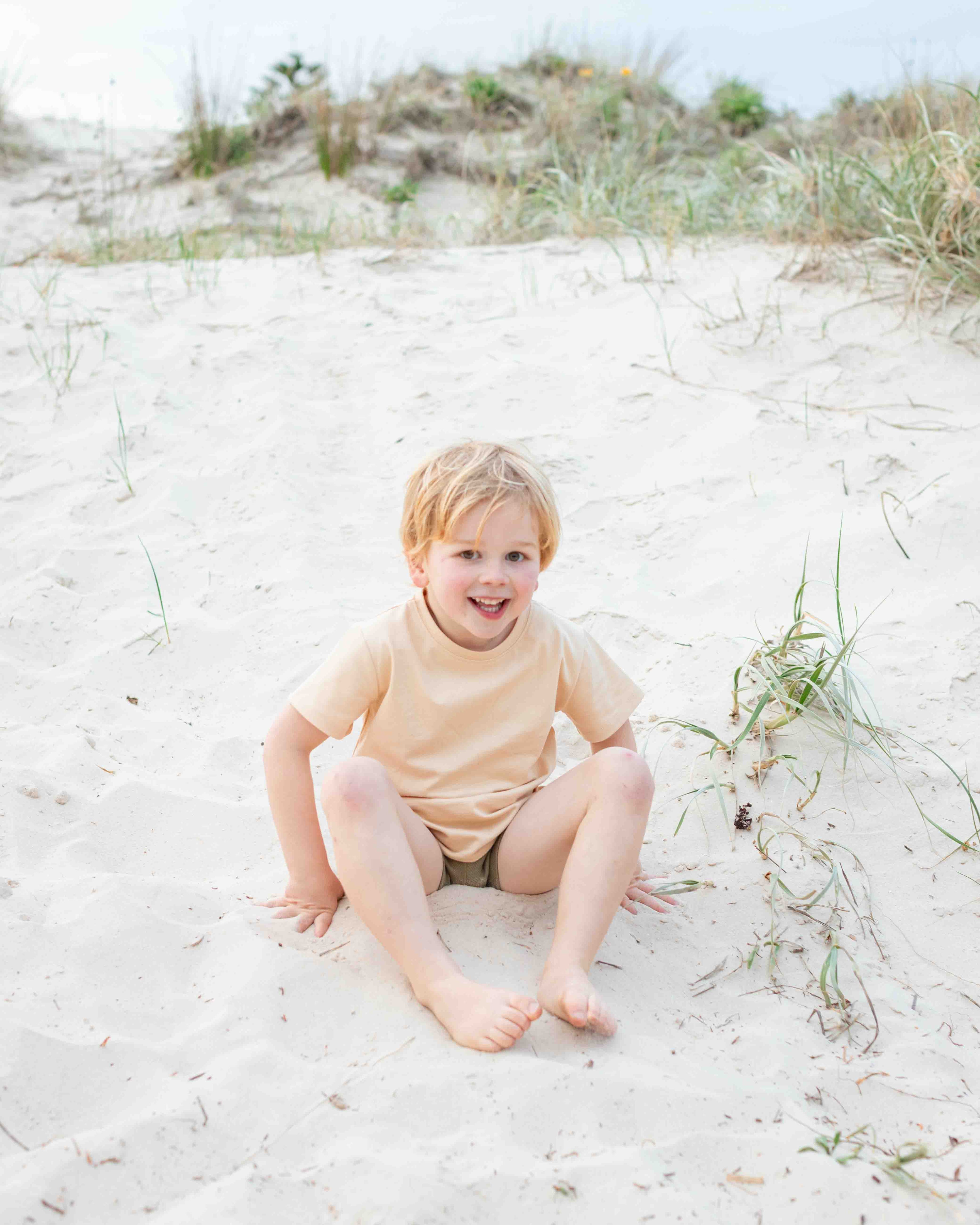 Child sitting on a sandy beach with grass and sand dunes in the background