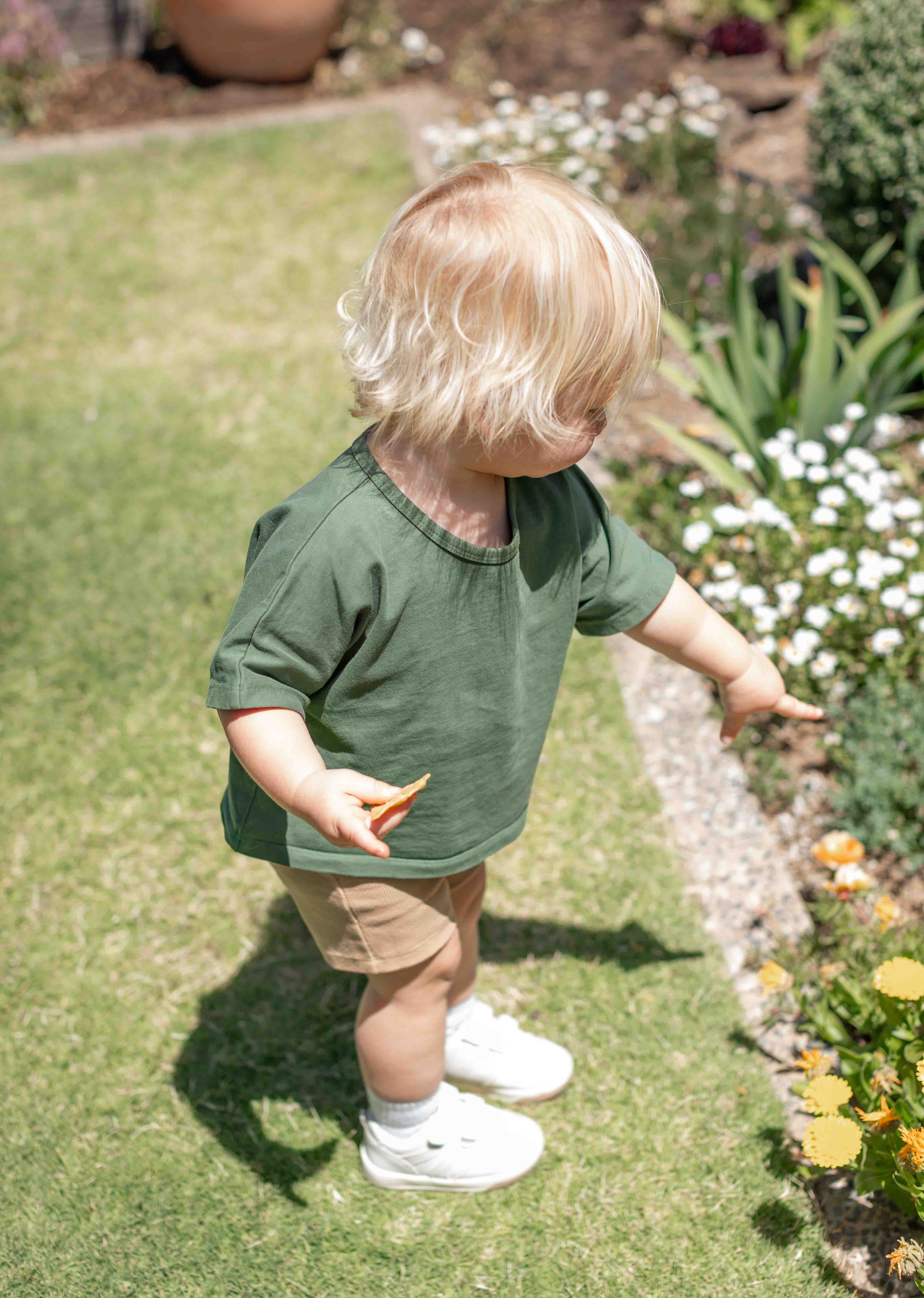 Child in a green shirt standing in a garden with flowers and plants.