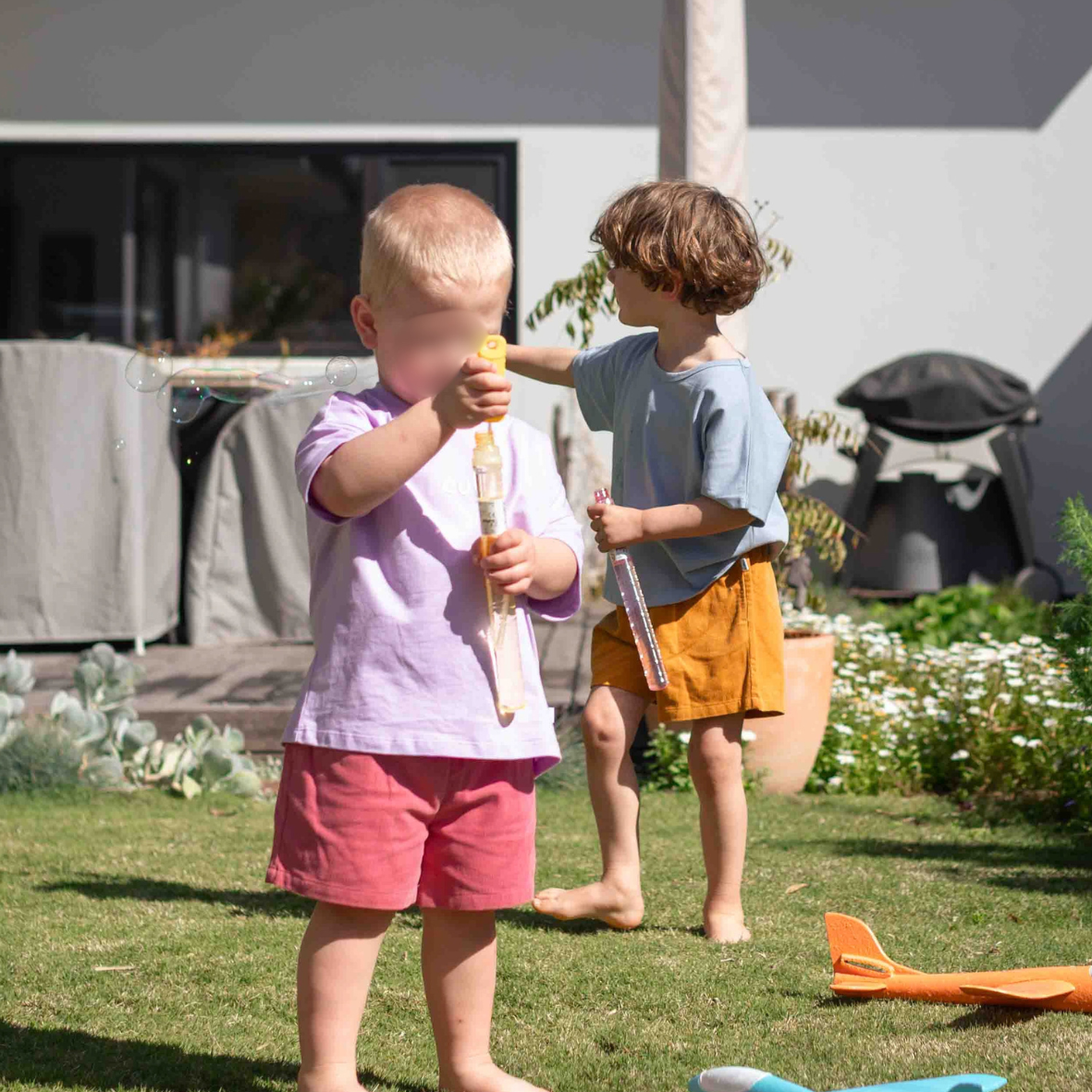 Two young children playing with bubble wands on a grassy backyard lawn. The child in the foreground wears a light purple t-shirt and pink shorts, while the child behind wears a light blue t-shirt and orange shorts.