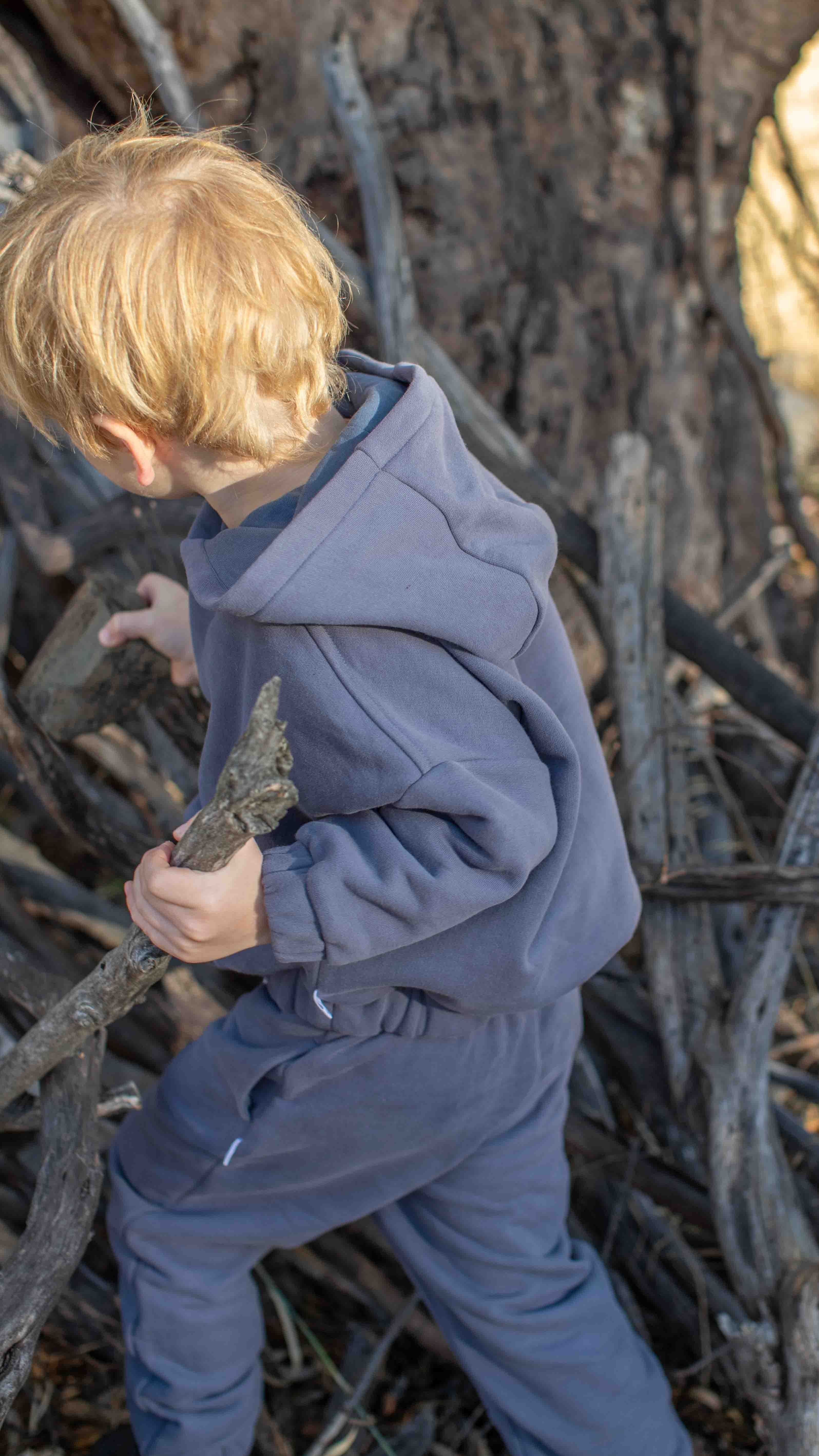 A young child with blond hair wearing a matching slate blue hooded sweatshirt and sweatpants tracks a twig through a forest of tree trunks and branches.