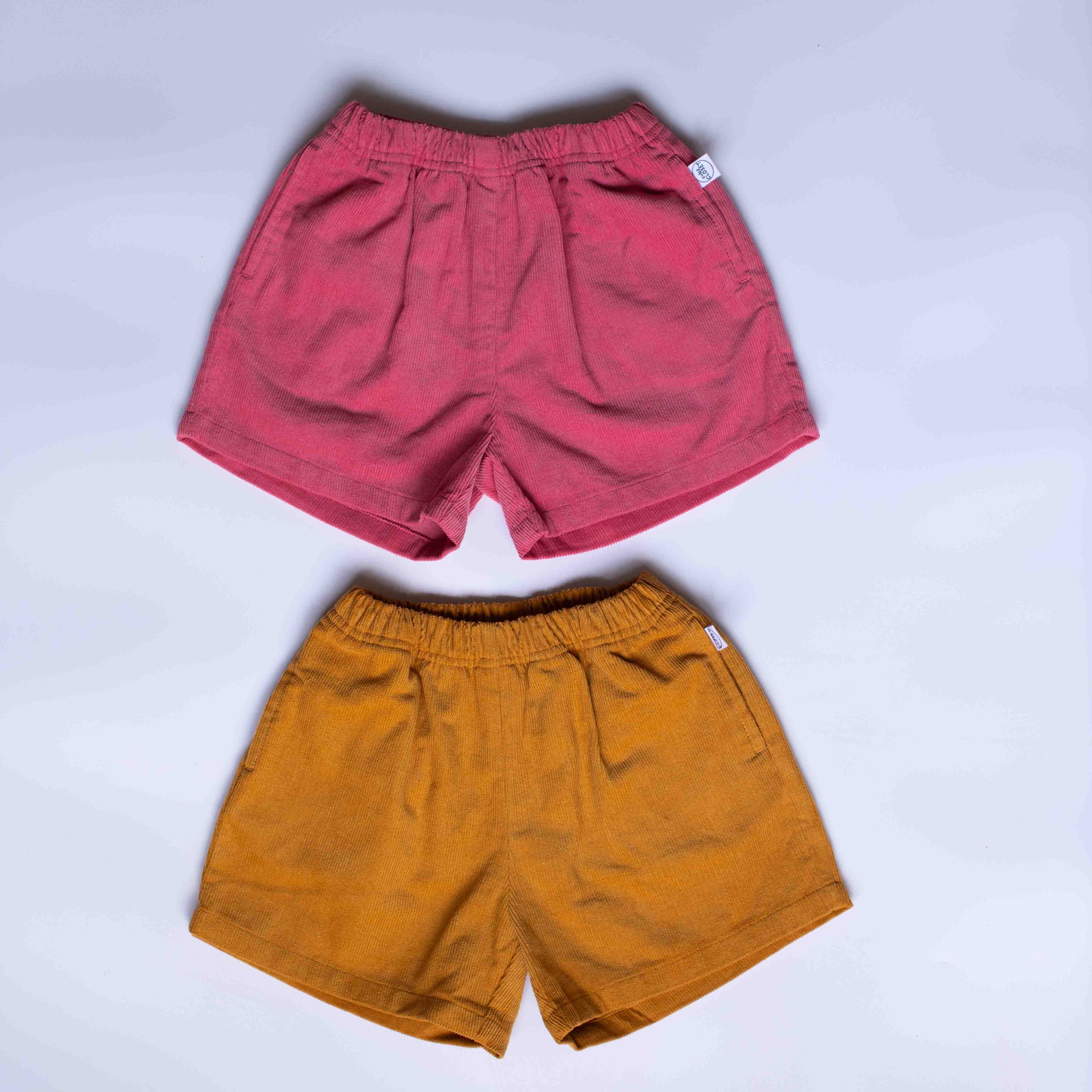 Two pairs of casual elastic-waist shorts laid flat on a light gray background. The top pair is dusty rose pink, and the bottom pair is mustard yellow.