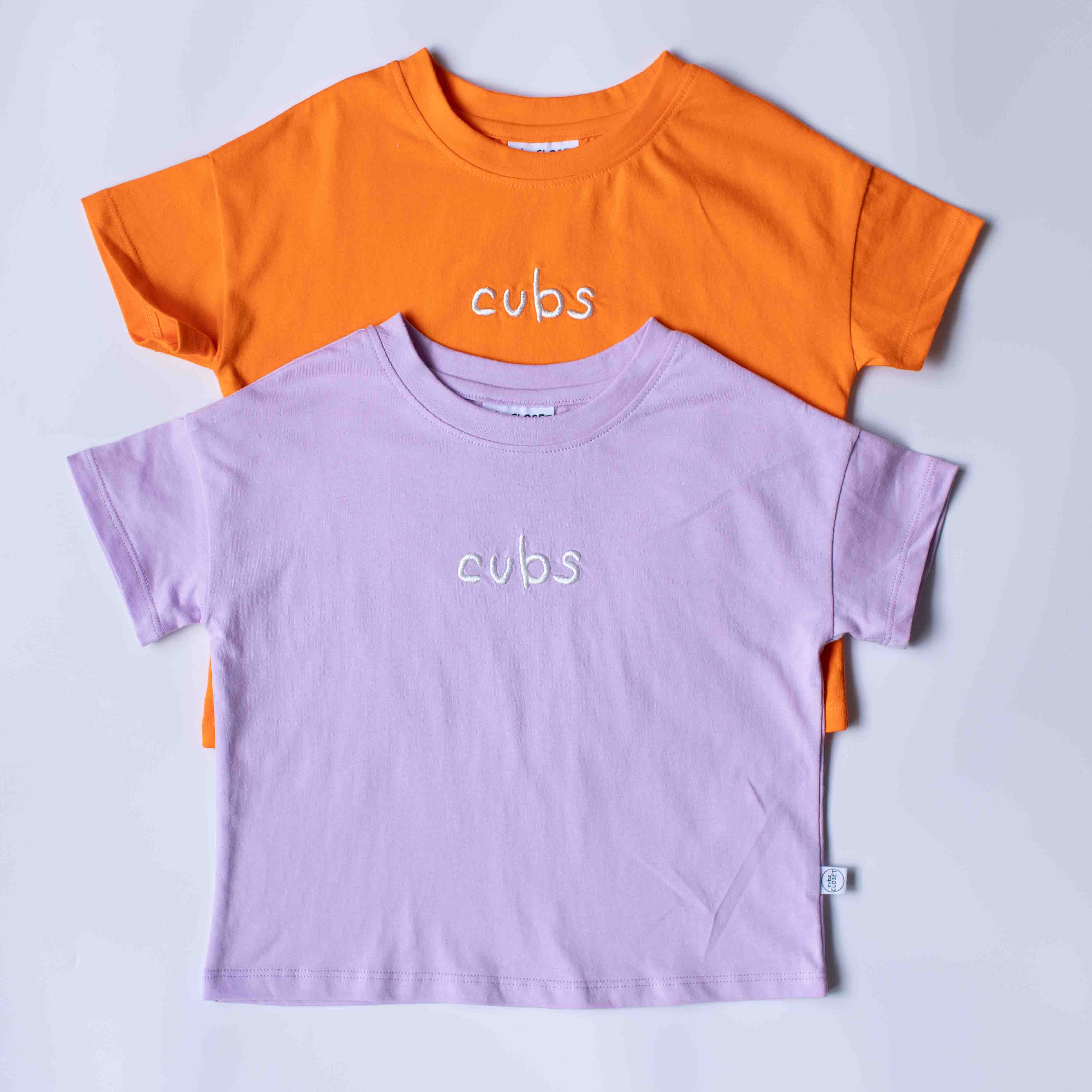 Two short-sleeve crewneck t-shirts for kids, one in orange and one in light purple, both featuring the word 'cubs' printed in white script across the chest, arranged in a flat-lay on a light gray background.