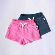 Two pairs of casual drawstring shorts stacked on a light gray background. The front pair is bright pink, and the pair behind it is dark teal.