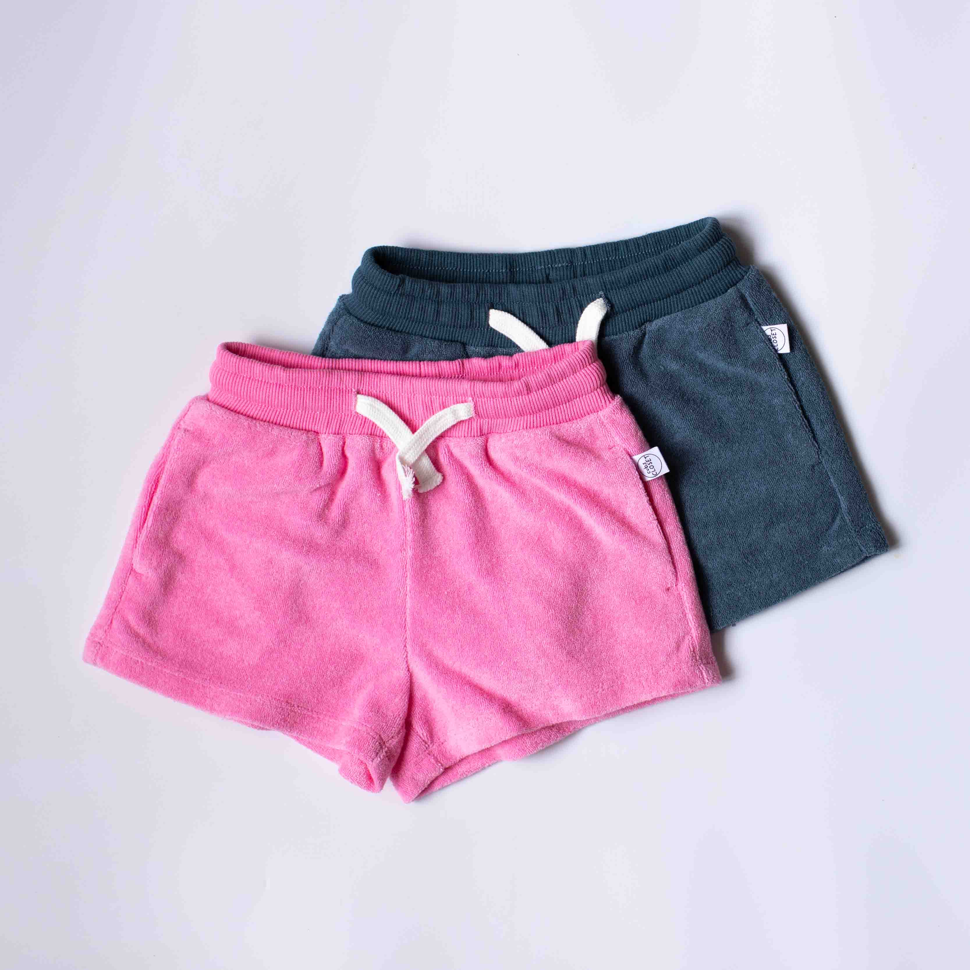 Two pairs of casual drawstring shorts stacked on a light gray background. The front pair is bright pink, and the pair behind it is dark teal.