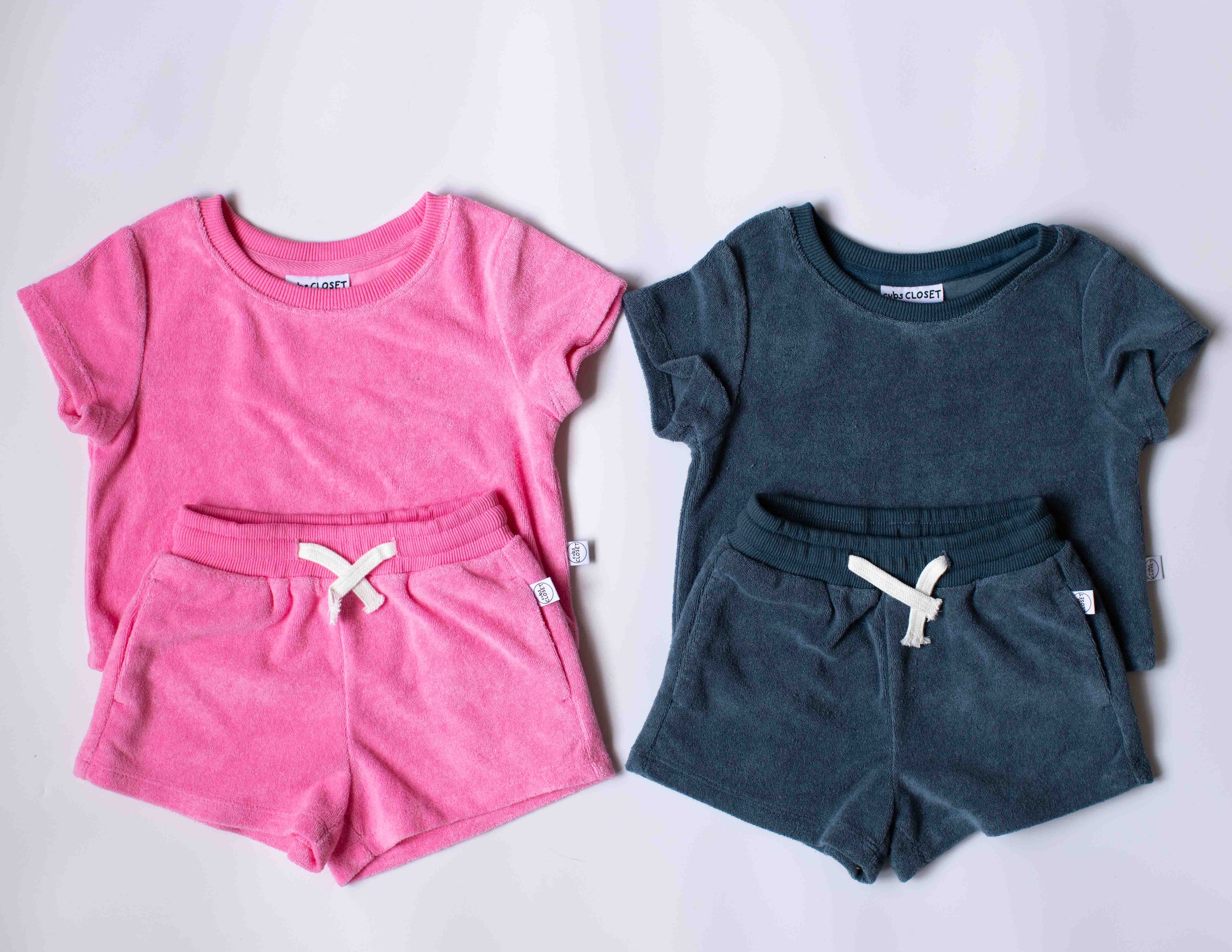 Two children's short-sleeve lounge sets laid flat on a light background. The left set is bright pink with white drawstrings, and the right set is dark teal with white drawstrings.
